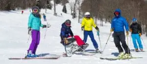 A woman on a sit ski is surrounded by standing skiers in bright ski gear.